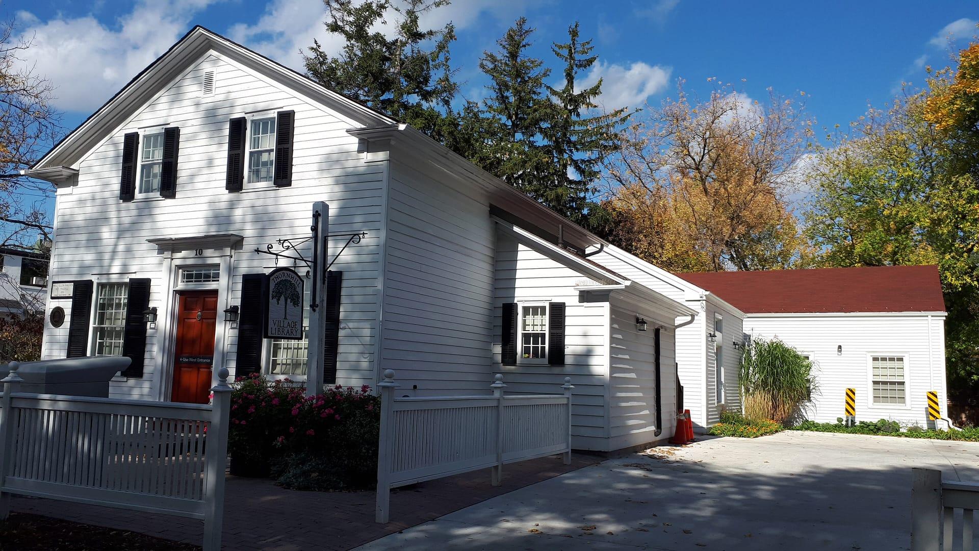 A white two-storey clapboard building with a red door, the Thornhill Village Library, in the fall